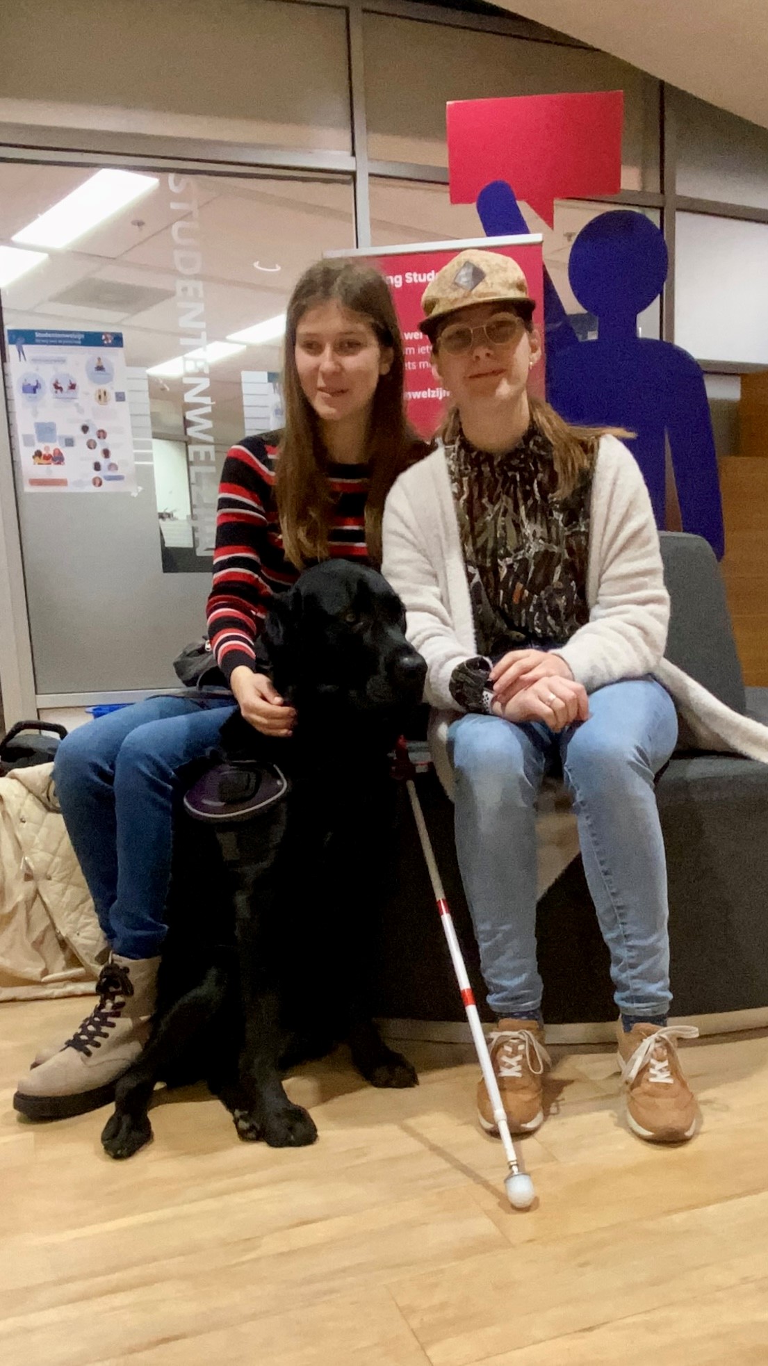 Claudia en Kim zitten naast elkaar. Links is Kim, een witte vrouw met bruin haar, Ze draagt een gekleurde trui en een spijkerbroek. Rechts zit Claudia, een witte vrouw, ze draagt een bril en een pet. Tussen hen in zit Kim’s hulphond, Fender.  